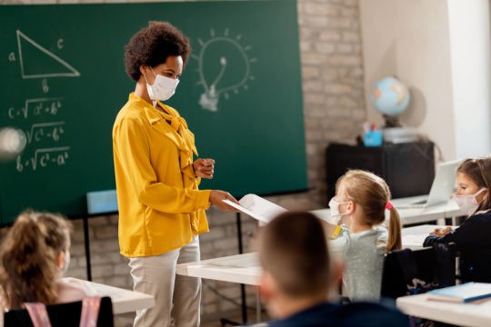 Masked teacher giving a paper to a masked student in a classroom.