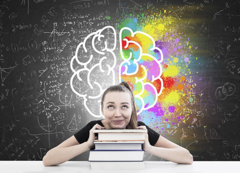 A girl sits resting her head on a stack of books while looking up at a brain sketch on the blackboard behind her.